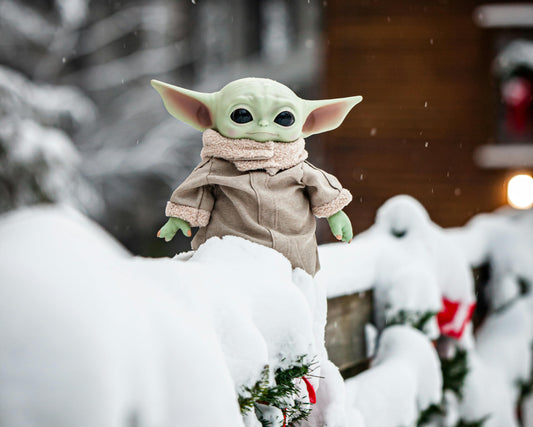 Plush toy resembling 'The Child' from 'The Mandalorian' on a snow-covered railing with a blurred snowy background.